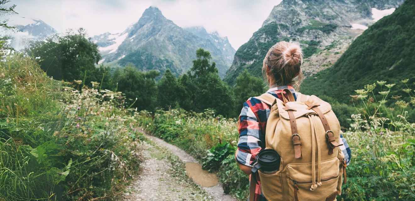 Žena s batohem stojící na cestě a hledí na vrcholky hor. Je na své misi splnit Mystery Hike výzvu.