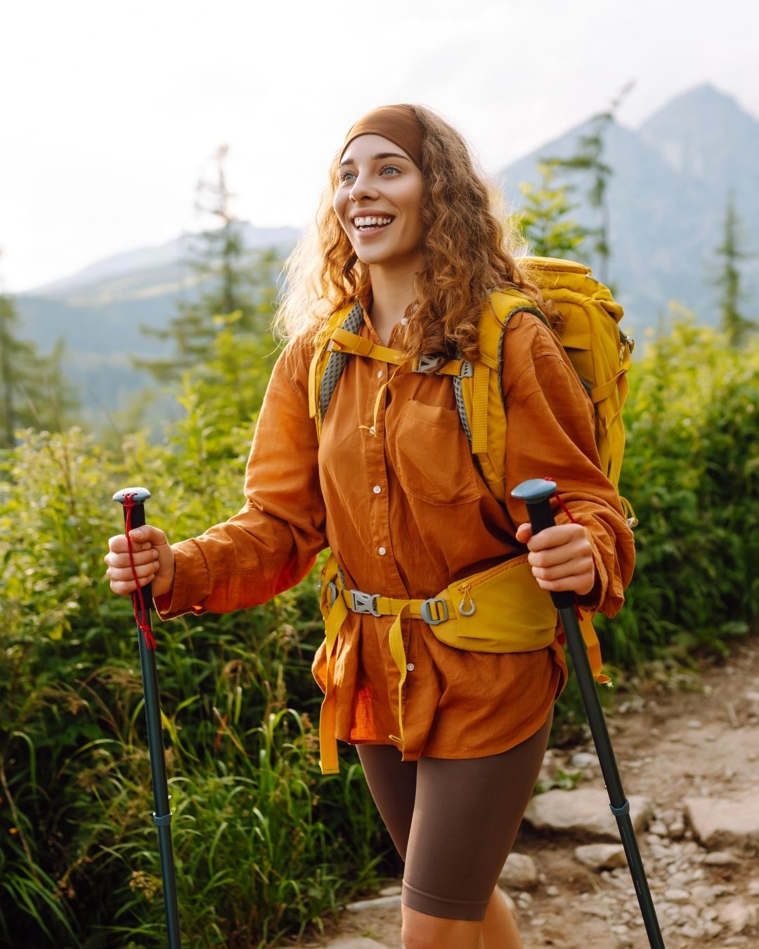 Žena na túře v přírodě. Zrzavé vlasy, oranžová košile, žlutý batoh a drží turistické hole.